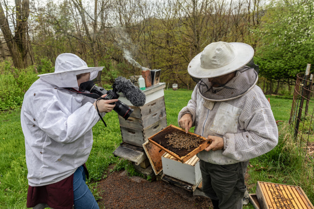 Sidney Beeman in a beekeeper suit film's beekeeper who is pointing at a hive with bees on it